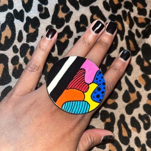 May include: A large, round ring with a colorful abstract design. The ring features black, white, pink, red, orange, blue, and yellow sections. The ring is worn on a hand with dark nail polish.