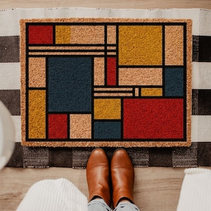 May include: A rectangular doormat with a geometric design in red, yellow, blue, and tan. The mat has a black border and is made of a textured material. Brown boots and blue jeans are visible in the foreground.