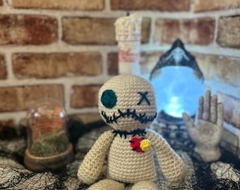 May include: A beige crochet doll with black stitched facial features, a teal eye, and a small heart-shaped embellishment. The doll is posed on a black web-like fabric, with a brick wall background and decorative items.