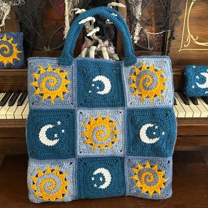 May include: A handmade crochet tote bag and matching square pouch. The bag features a repeating pattern of yellow suns, white moons, and stars on a blue and gray background. The bag has a blue handle.