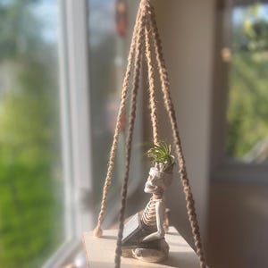 Peut inclure: Une étagère suspendue en bois avec une petite figurine de squelette tenant un livre et une plante dans sa tête. L'étagère est suspendue par une corde de jute et des perles de bois. La figurine est blanche et la plante a des feuilles vertes.