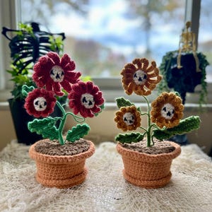 May include: Two crochet flower arrangements in terracotta pots. One has red flowers with white skull centers, the other has orange flowers with white skull centers. Green leaves and stems complete the floral design. The pots are sitting on a white surface.