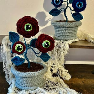 May include: Crocheted eye-flower sculptures in light blue pots. One features burgundy flowers with green and black centers, the other has blue flowers. The stems are teal, and the leaves are blue. The pots are sitting on a wooden surface.