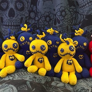 Könnte beinhalten: Eine Sammlung handgefertigter Häkelpuppen in verschiedenen Farben. Die Puppen umfassen schwarze, blaue, gelbe und rote Figuren mit aufgestickten Gesichtszügen und herz- oder M-förmigen Details. Der Hintergrund zeigt ein Totenkopfmuster.