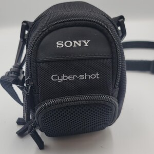 May include: A black camera case with a textured fabric exterior, featuring the white text SONY and Cyber-shot on the front. The case includes a zippered main compartment, a smaller front pocket with a mesh panel, and an attached black shoulder strap.