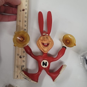 Peut inclure: Une figurine de lapin de dessin animé rouge et beige, tenant deux objets en plastique jaune. La figurine porte un "N" blanc sur la poitrine et mesure environ 20 cm de haut.