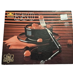 May include: A Golden 550-piece jigsaw puzzle featuring a still life of cherries in a black bowl with a fork. The puzzle box has a black and orange striped background. The puzzle is fully interlocking.