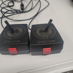 May include: Two black Radio Shack joysticks with red buttons and a central joystick. The joysticks have a circular design with a directional arrow. Black cords and a connector are visible in the background.
