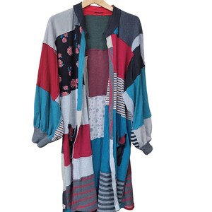 May include: A long-sleeved cardigan featuring a patchwork design with various fabric panels in red, teal, grey, and black. The garment includes sections with floral patterns, horizontal stripes, and solid colors, finished with ribbed cuffs and a collar.