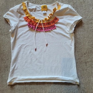 Peut inclure: Un t-shirt blanc à manches courtes doté d'une encolure ronde ornée de volants en tissu superposés dans des tons jaune, orange et rose. L'encolure comprend un cordon de serrage blanc avec de petites perles décoratives aux extrémités et une étiquette.