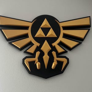 以下が含まれることがあります： ゼルダの伝説のハイリア紋章をあしらった、黒と金の壁飾り。エンブレムは黒い背景に金色の翼と中央に金色のトライフォースのシンボルがあります。デザインは白い壁に取り付けられています。