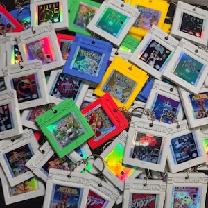 May include: A collection of colorful Game Boy cartridge keychains. The keychains feature various game titles like "Pokemon", "Alien", and "Star Wars". The cartridges are in various colors, including red, green, blue, and yellow.