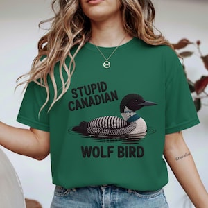 Pode incluir: T-shirt verde com um gráfico de um mergulhão e o texto "STUPID CANADIAN WOLF BIRD". O pássaro é preto e branco com um pescoço azul e branco. O texto é preto.