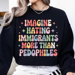 Op de afbeelding: Zwart sweatshirt met de tekst "IMAGINE HATING IMMIGRANTS MORE THAN PEDOPHILES" in kleurrijke retro-letters. De tekst is gestapeld met steraccenten. Een casual kledingstuk.