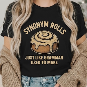 May include: Black t-shirt featuring a cinnamon roll graphic and the text "SYNONYM ROLLS JUST LIKE GRAMMAR USED TO MAKE". The design is in tan and brown tones, depicting a detailed illustration of a baked pastry.