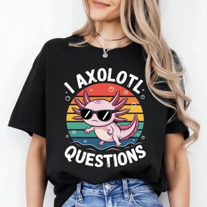 May include: Black t-shirt featuring a pink axolotl wearing sunglasses, set against a colorful sunset design. The text "I AXOLOTL QUESTIONS" is printed above and below the axolotl illustration. The shirt is made of a soft material.