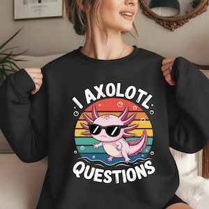 May include: Black sweatshirt featuring a pink axolotl wearing sunglasses, set against a colorful sunset design. The text "I AXOLOTL QUESTIONS" is printed in white. A fun, casual top.