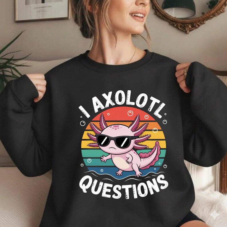 May include: Black sweatshirt featuring a pink axolotl wearing sunglasses, set against a colorful sunset design. The text "I AXOLOTL QUESTIONS" is printed in white. A fun, casual top.