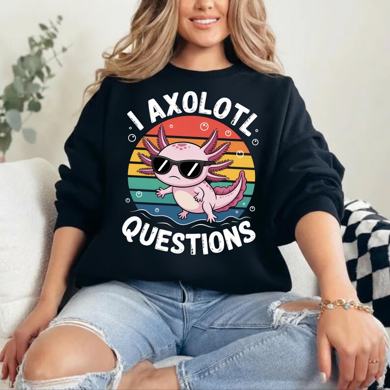 May include: Black sweatshirt featuring a colorful graphic of a pink axolotl wearing sunglasses, set against a sunset background. The text "I AXOLOTL QUESTIONS" is printed in white. The sweatshirt is a casual, long-sleeved top.