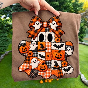 Pode incluir: Uma t-shirt castanha com uma grande aplicação central em forma de fantasma. O fantasma é composto por um patchwork de padrões xadrez laranja, pretos e brancos, decorado com fantasmas, abóboras, morcegos e chapéus bordados.