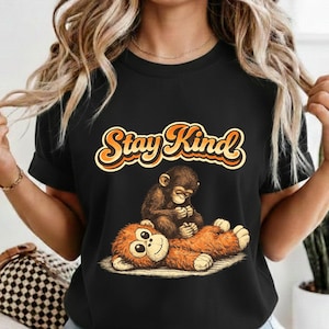 May include: Black t-shirt with the text "Stay Kind" in orange and yellow script. The shirt features a cartoon illustration of a small monkey comforting a larger, orange monkey. The design is centered on the shirt.