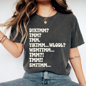 Puede incluir: Camiseta gris oscuro con texto blanco. El texto incluye frases como "DYKTMM?" y "TMM?" en una fuente informal y ligeramente desgastada. La camiseta tiene un corte holgado, adecuada para el uso diario.