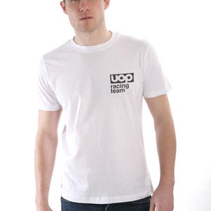Puede incluir: Camiseta blanca de cuello redondo con el texto "uoq racing team" en un cuadrado negro. El modelo lleva vaqueros azules. La camiseta es de manga corta y está hecha de un material suave.