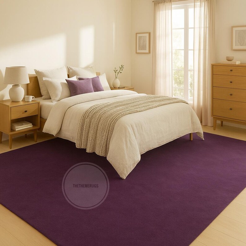 May include: A bedroom with a deep purple rug covering the floor. The rug is under a bed with white linen and purple cushions. A wooden bedside table with a lamp and a chest of drawers are also visible. The room has a neutral colour scheme.