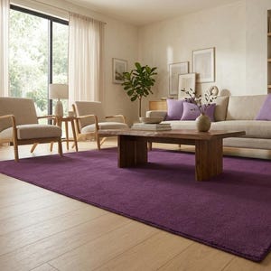 May include: A living room with a deep purple rectangular rug. The room features a wooden coffee table, a beige sofa with purple cushions, and two beige armchairs. Natural light enters through a large window.