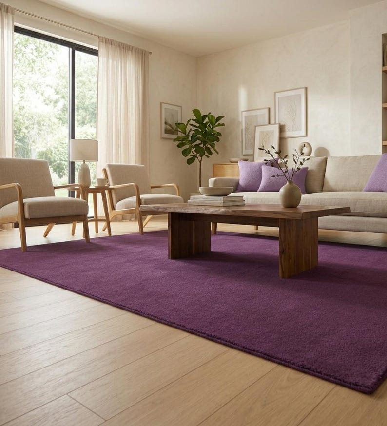 May include: A living room with a deep purple rectangular rug. The room features a wooden coffee table, a beige sofa with purple cushions, and two beige armchairs. Natural light enters through a large window.