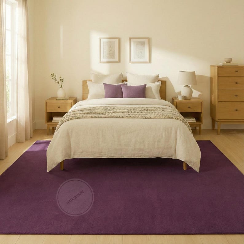 May include: A bedroom with a deep purple rug. The rug is beneath a bed with a light-coloured wooden frame and matching bedside tables. The bed has white bedding and purple cushions. A wooden chest of drawers is on the right.