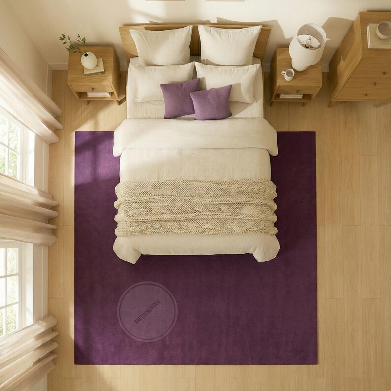 May include: An overhead shot of a bedroom featuring a deep purple rug. A bed with white bedding, two purple cushions, and a beige throw is the focal point. Two wooden bedside tables are either side of the bed, and a window with blinds is visible.