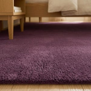 May include: A close-up of a deep purple rug with a soft, textured pile. The rug is placed on a light-coloured wooden floor, near a wooden bed frame and a bedside table. The rug's colour is a rich, dark shade.