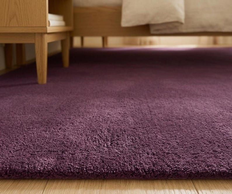 May include: A close-up of a deep purple rug with a soft, textured pile. The rug is placed on a light-coloured wooden floor, near a wooden bed frame and a bedside table. The rug's colour is a rich, dark shade.