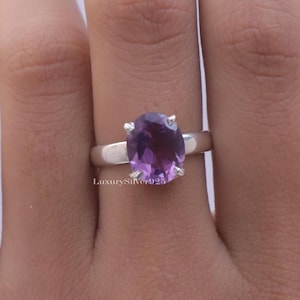 Op de afbeelding: Een zilveren ring met een grote, ovaal geslepen paarse edelsteen in een zetting met vier pootjes. De ringband is van glad, gepolijst zilver. De tekst LuxurySilver925 is zichtbaar op de ring, wat de metaalsamenstelling van het sieraad aangeeft.