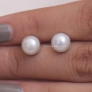 Könnte beinhalten: Ein Paar runde, weiße Perlenohrstecker mit einer silbernen Lünettenfassung. Die Ohrringe werden auf hellem Hintergrund präsentiert. Der Text "LuxurySilver925" ist sichtbar.