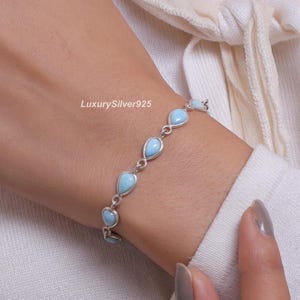 May include: A silver chain bracelet featuring multiple teardrop-shaped light blue stones set in metal bezels. The bracelet is delicate and links the stones together in a repeating pattern. The text LuxurySilver925 is visible near the jewellery.