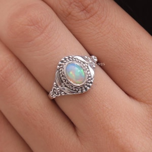 May include: A sterling silver ring featuring an oval-shaped iridescent opal gemstone set in a decorative bezel. The band displays intricate metalwork with beaded detailing and leaf-like patterns surrounding the central stone, stamped with 925.