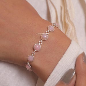 Könnte beinhalten: Ein silbernes Kettenarmband mit einer Reihe runder, glatter, hellrosa Cabochon-Steine, die in polierte Silberfassungen eingefasst sind. Das Armband besteht aus einzelnen kreisförmigen Gliedern, die jede Steinfassung in einem durchgehenden Design verbinden.