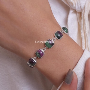 May include: A silver link bracelet featuring five square-cut, polished ruby zoisite gemstones. Each stone displays a mix of deep red and forest green hues, set in individual silver bezels connected by small silver rings. The text LuxurySilver925 is visible.