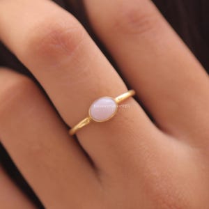 Könnte beinhalten: Ein dünner Goldring mit einem kleinen, glatten, ovalen hellrosa Stein, der horizontal in der Mitte gefasst ist. Der Ring hat eine polierte metallische Oberfläche und ein schlichtes, zartes Design, das mittig am Finger sitzt.