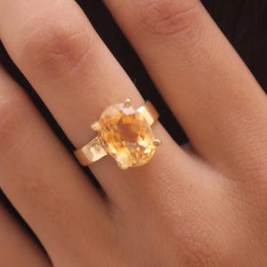Peut inclure: Une bague dorée ornée d'une grande pierre précieuse jaune translucide de taille ovale, montée sur un anneau métallique simple. La pierre est facettée et reflète la lumière, tandis que l'anneau présente une finition lisse et polie. Le texte LuxurySilver est visible.