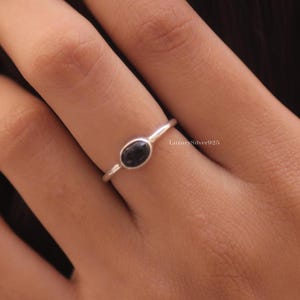 May include: A thin silver ring featuring a small, smooth, oval-shaped black stone set in a simple metal bezel. The band is slender and polished, showcasing a minimalist design with the text LuxurySilver925 visible near the setting on the band.