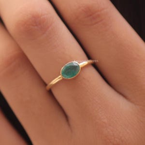 May include: A gold ring featuring a small, oval-shaped green gemstone set in a bezel setting. The band is thin and polished, with the text LuxurySilver925 visible in small white letters near the setting of the ring on the finger.
