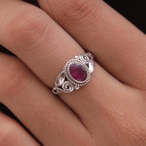 May include: A silver ring featuring an oval, deep red gemstone. The ring has an ornate design with swirling patterns and small beaded accents around the gemstone. The ring is shown on a finger.