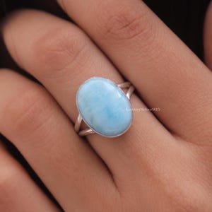 May include: A sterling silver ring featuring a large, oval-shaped, light blue Larimar gemstone with a smooth, polished surface. The band is split into two thin, polished silver wires that meet at the base of the stone setting. Text reads LuxurySilver925.