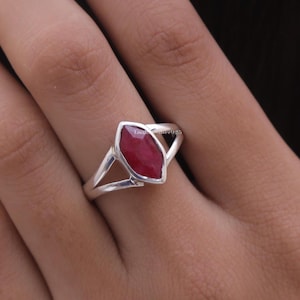 Op de afbeelding: Een zilveren ring met een marquise-geslepen, dieprode edelsteen in een zetting. De ringband heeft een gespleten ontwerp dat taps toeloopt naar de steen. De tekst "LaxmiSilver925" is vaag zichtbaar aan de zijkant van de zilveren zetting.