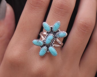 Puede incluir: Un anillo de plata con un grupo de siete piedras de turquesa de forma ovalada dispuestas en un patrón floral. Cada piedra está engastada en un bisel de plata dentado y la banda del anillo se compone de varios hilos de plata pulida.