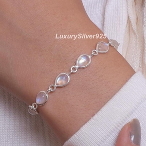 Pode incluir: Uma pulseira de corrente de prata com cinco cabochões de pedra da lua em forma de lágrima ligados entre si. A pulseira apresenta o texto LuxurySilver925. As pedras exibem uma qualidade translúcida e iridescente em engastes de prata polida.