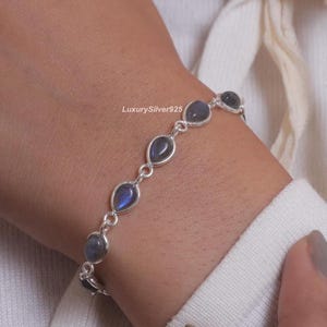 Peut inclure: Un bracelet en chaîne argentée composé de plusieurs pierres en forme de goutte d'eau aux reflets bleu-gris. Les maillons sont en argent poli et le texte LuxurySilver925 est visible près du centre du design du bracelet.
